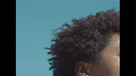 Profile view of a dark-skinned person's head and shoulders with curly afro against a blue sky.  Earrings are visible.