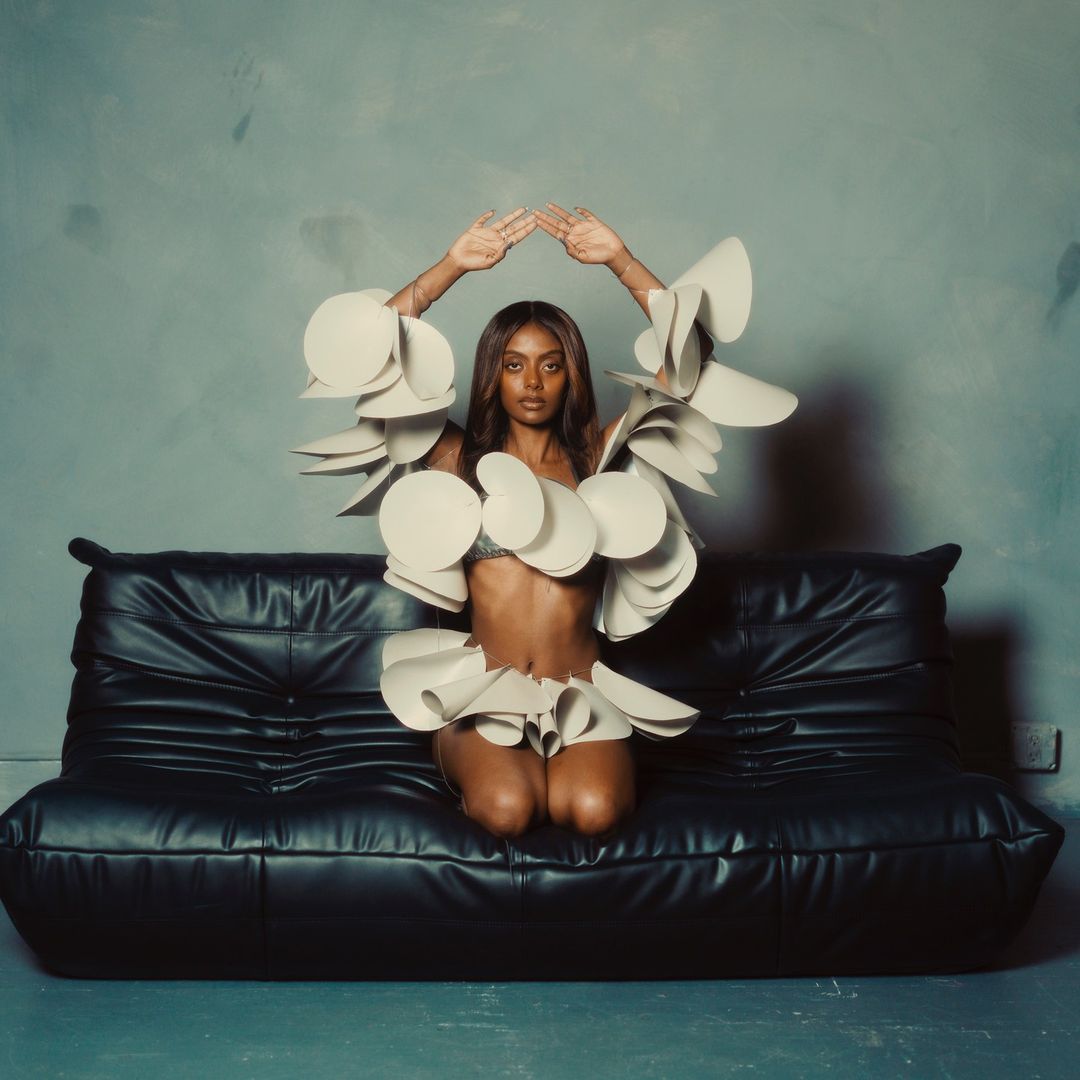 Woman in avant-garde fashion kneeling on a black leather sofa against a textured grey wall, hands raised overhead, studio lighting.