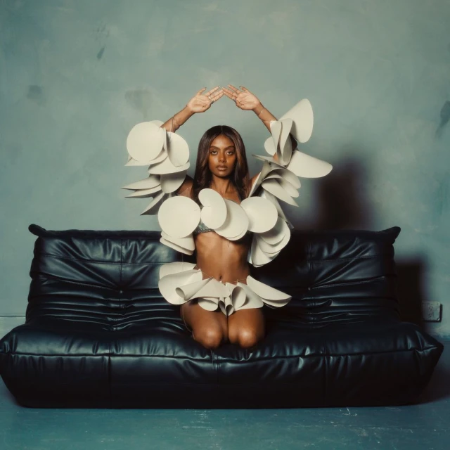 Woman in avant-garde fashion kneeling on a black leather sofa against a textured grey wall, hands raised overhead, studio lighting.