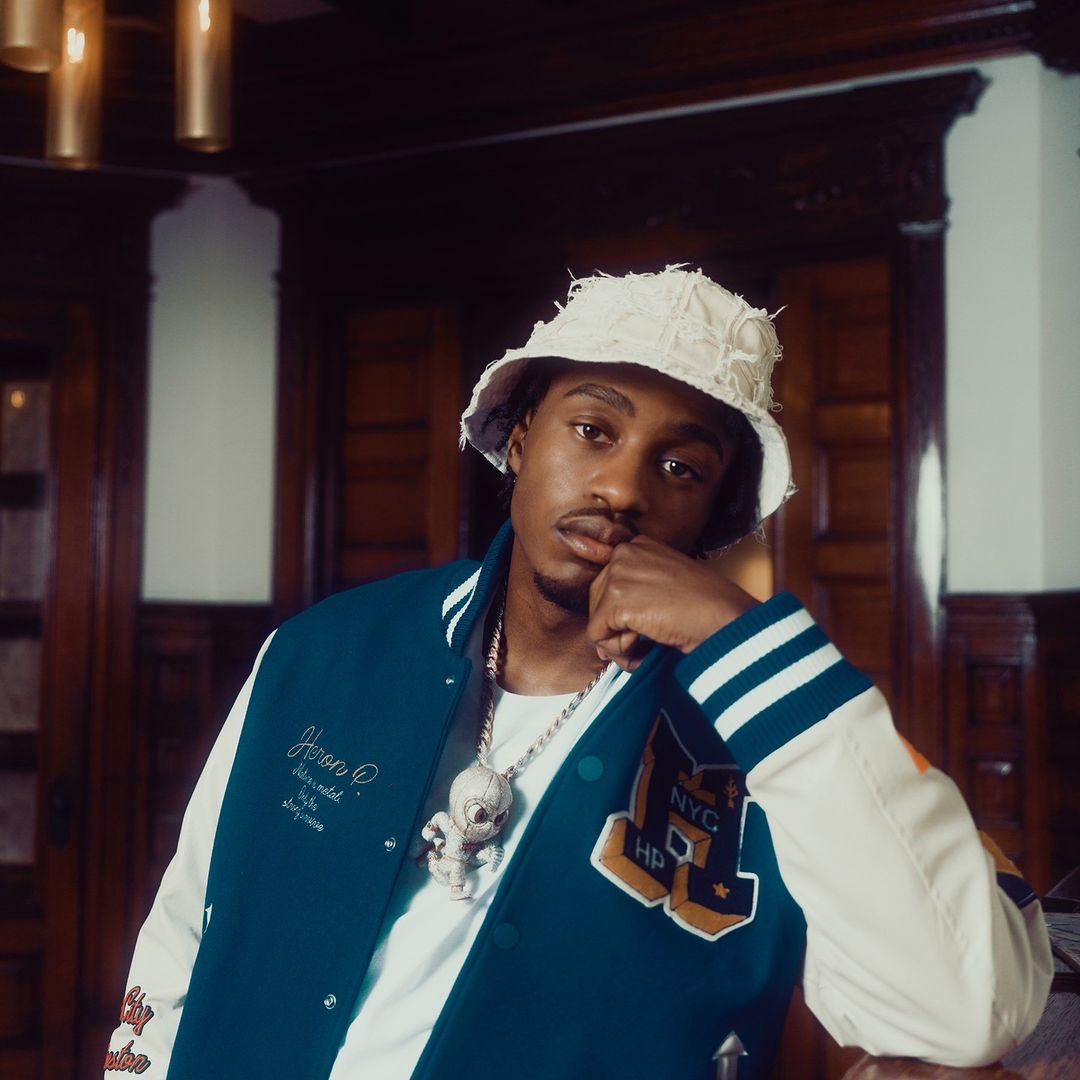 Adult male with a bucket hat and varsity jacket poses in a room with dark wood paneling and warm lighting, showcasing an urban fashion style.