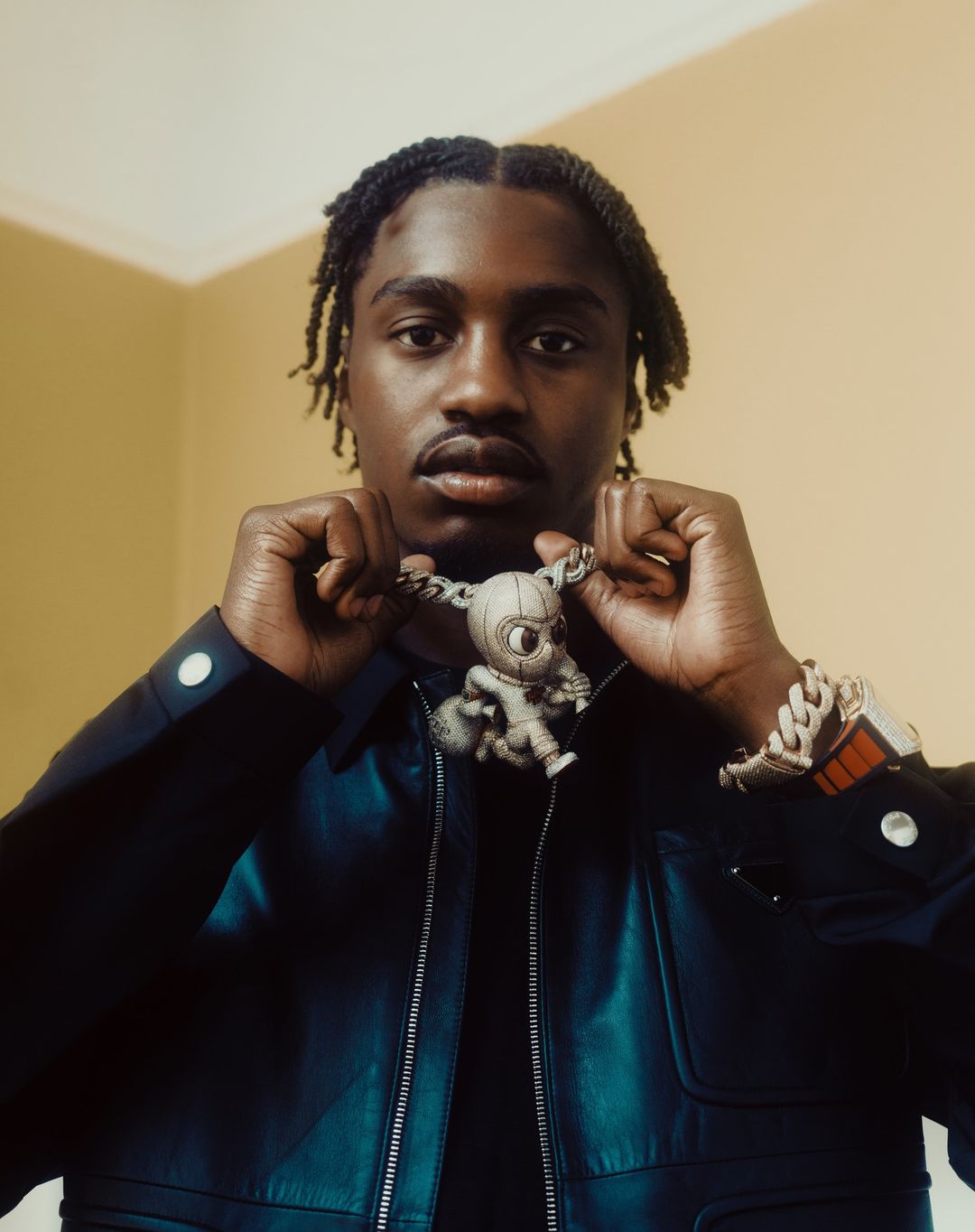 Headshot of an adult man with dreadlocks wearing a blue jacket and jewelry including an animated character pendant necklace.