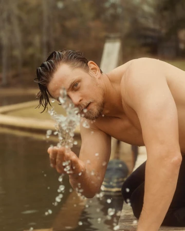 Shirtless man kneeling by water splashing droplets upward with his hand outdoors