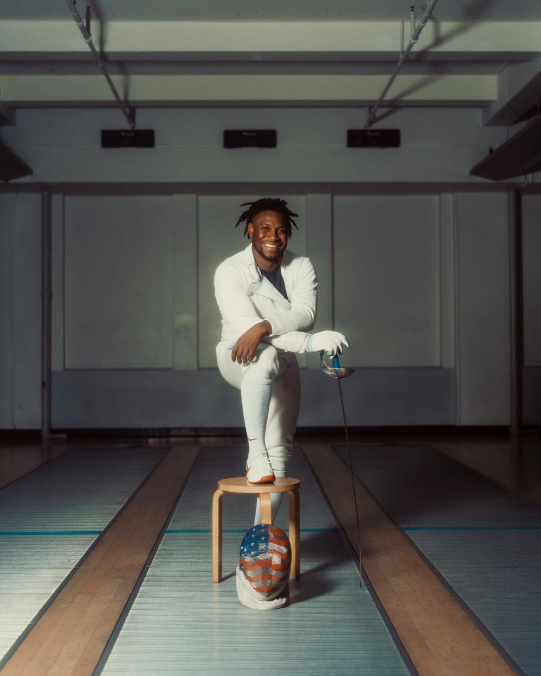 A smiling Black adult male fencer with dreadlocks poses in uniform with a foil, sitting on a stool, with a mask at the base.