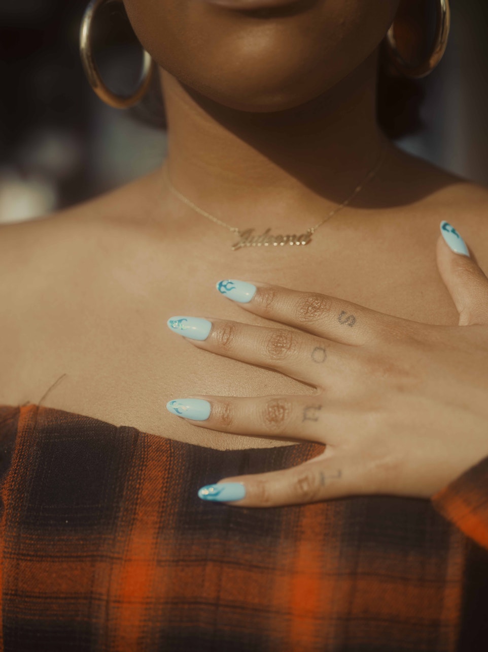 Close-up of a woman's chest and hand with painted nails. She wears gold hoop earrings and a necklace with a name pendant.