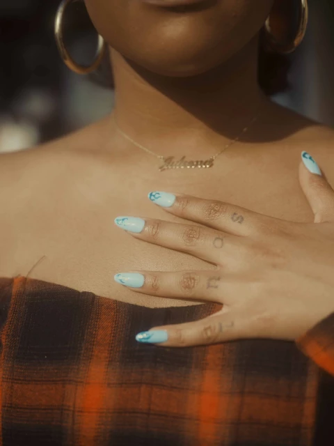Close-up of a woman's chest and hand with painted nails. She wears gold hoop earrings and a necklace with a name pendant.