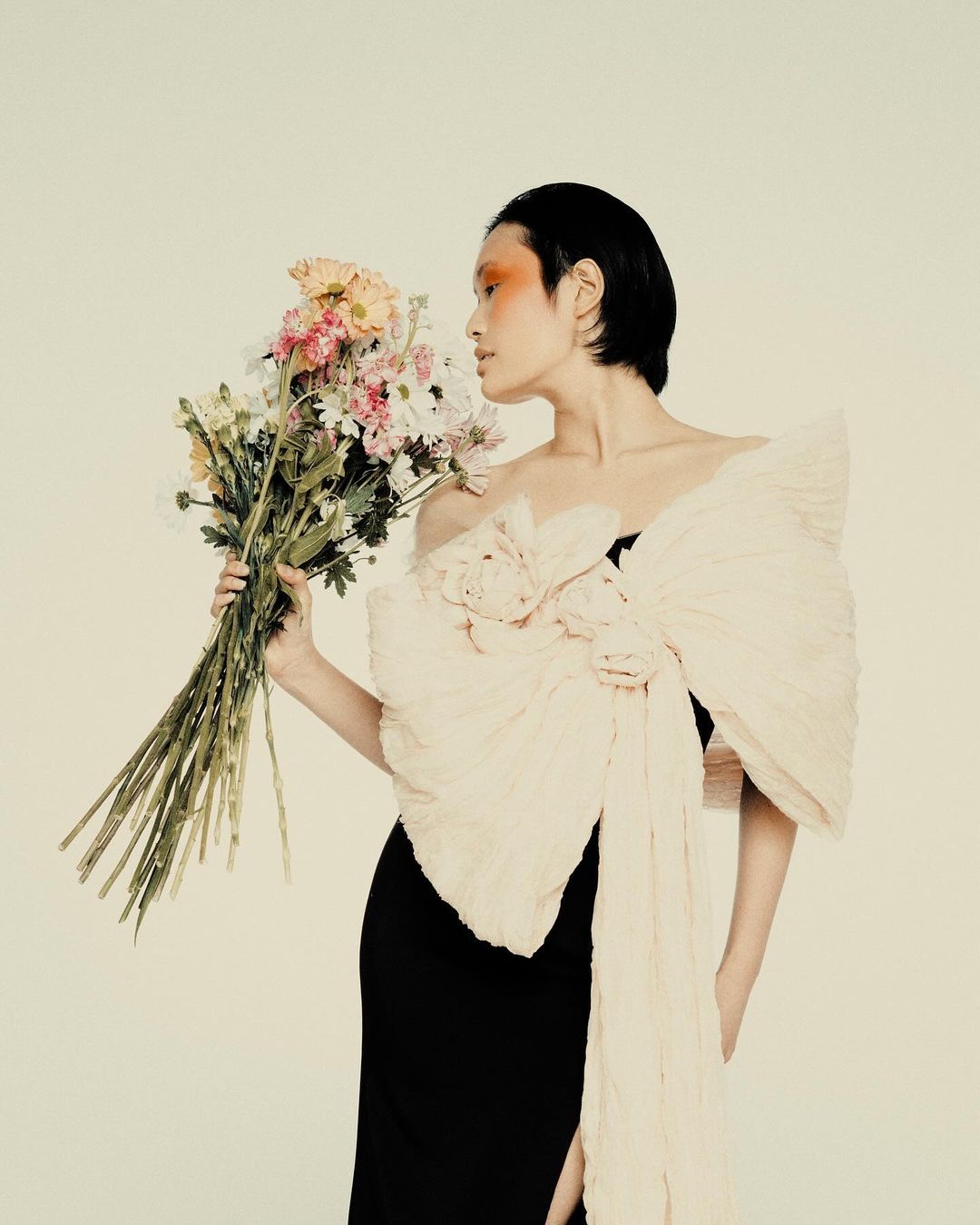 Elegant woman in a black dress poses with a bouquet of pastel flowers against a neutral backdrop in a fashion photography style studio shot.
