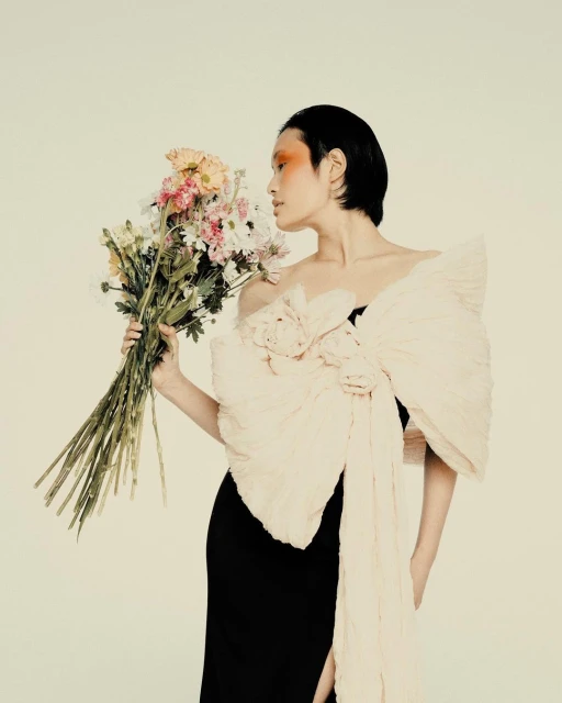 Elegant woman in a black dress poses with a bouquet of pastel flowers against a neutral backdrop in a fashion photography style studio shot.