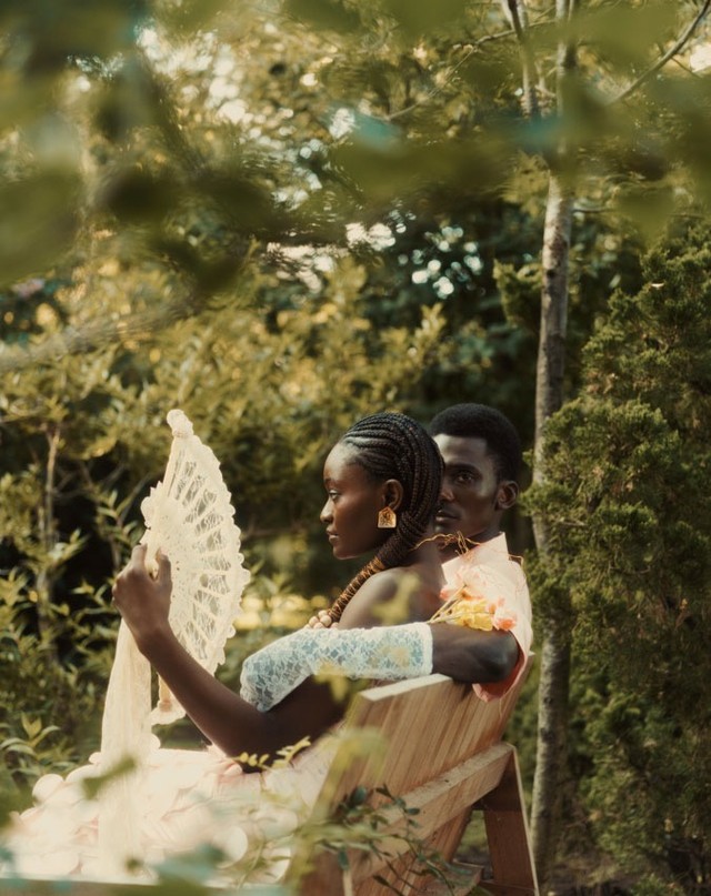 African American couple sitting on a bench in the garden. Woman is holding a fan. Man has his arm around the woman. 
