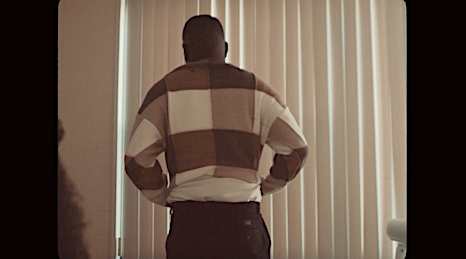 Adult male standing indoors near window in a patterned sweater. Casual fashion shot with soft lighting and simple background.