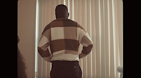 Adult male standing indoors near window in a patterned sweater. Casual fashion shot with soft lighting and simple background.