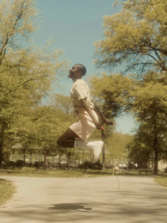 Athletic, smiling adult person jumping rope in a park setting on a sunny day with grass and trees in the background, wearing athletic clothing.