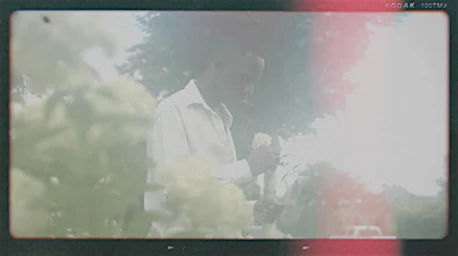 Adult male in white shirt with white flowers outdoors in bright sunlight, filmed with a vintage lens, resulting in light leaks and soft focus.
