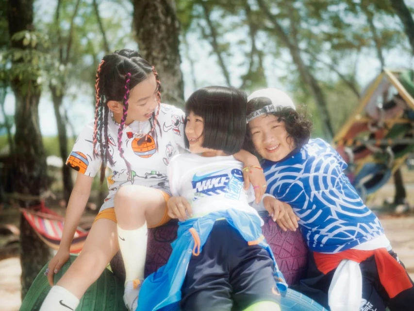 Three smiling Asian children wear colorful Nike shirts and accessories while sitting on a tire in a park-like outdoor setting.