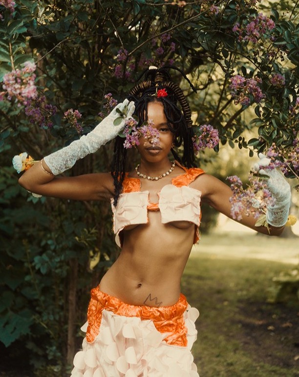 A woman in a creative outfit poses with outstretched arms in a garden setting, surrounded by purple flowers and green foliage.
