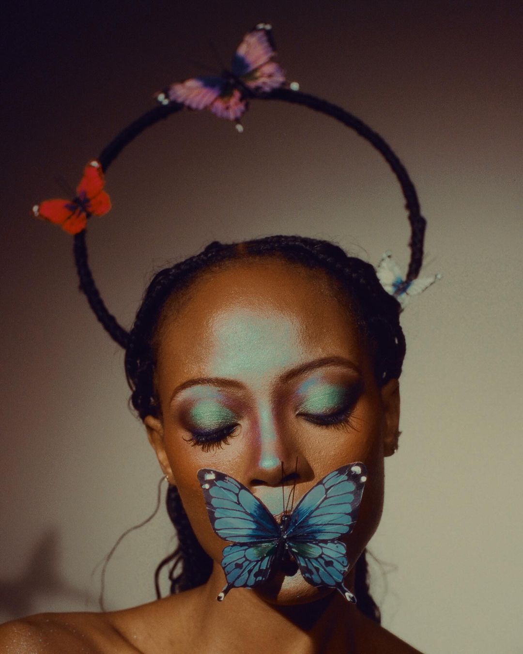 Close-up portrait of an adult female with blue eyeshadow and a butterfly attached to her lips, wearing a butterfly halo.