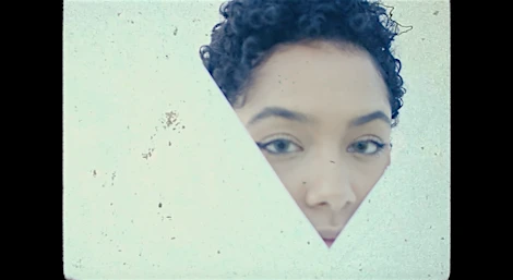 Close-up of a woman with short curly hair, peering through a triangular cutout in a textured, light blue surface with a film-grain effect.