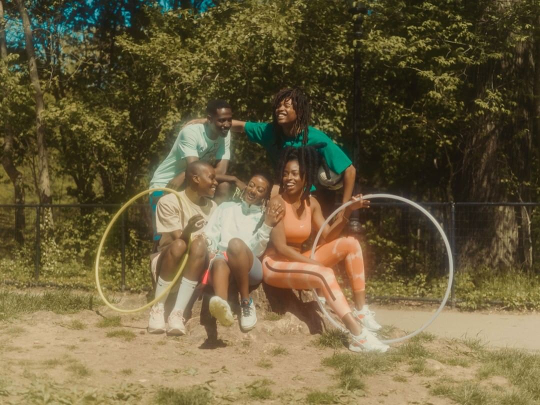 Group of five joyful people outdoors, sitting and standing together in a grassy park setting, holding hula hoops with trees behind them.