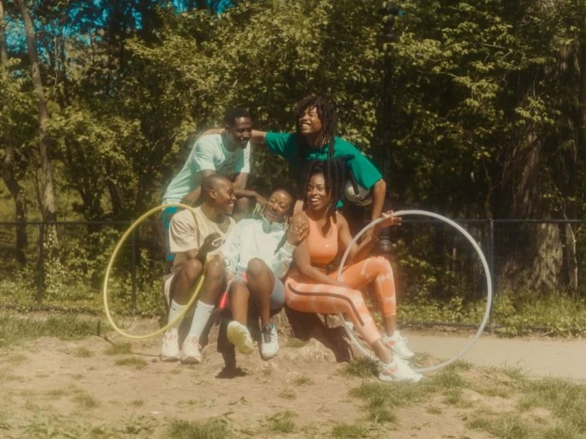 Group of five joyful people outdoors, sitting and standing together in a grassy park setting, holding hula hoops with trees behind them.