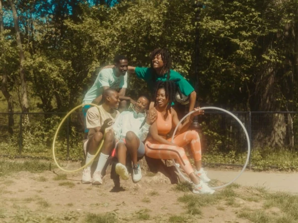 Group of five joyful people outdoors, sitting and standing together in a grassy park setting, holding hula hoops with trees behind them.