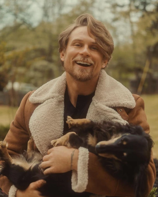 Happy man smiles while holding a black and brown dog outside. The man is wearing a sherpa jacket in an outdoor setting.