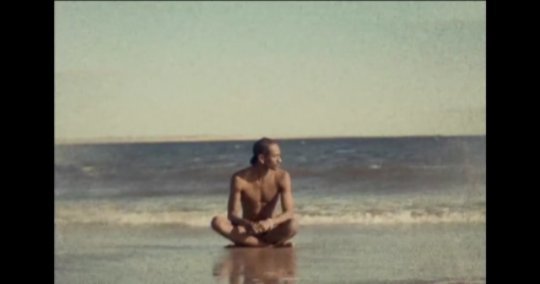 Man sitting cross-legged on a sandy beach near the ocean shoreline under an open sky
