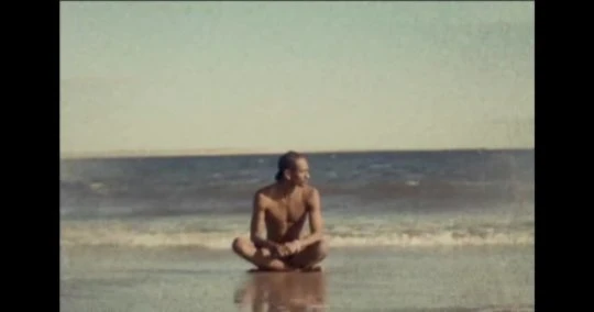 Man sitting cross-legged on a sandy beach near the ocean shoreline under an open sky