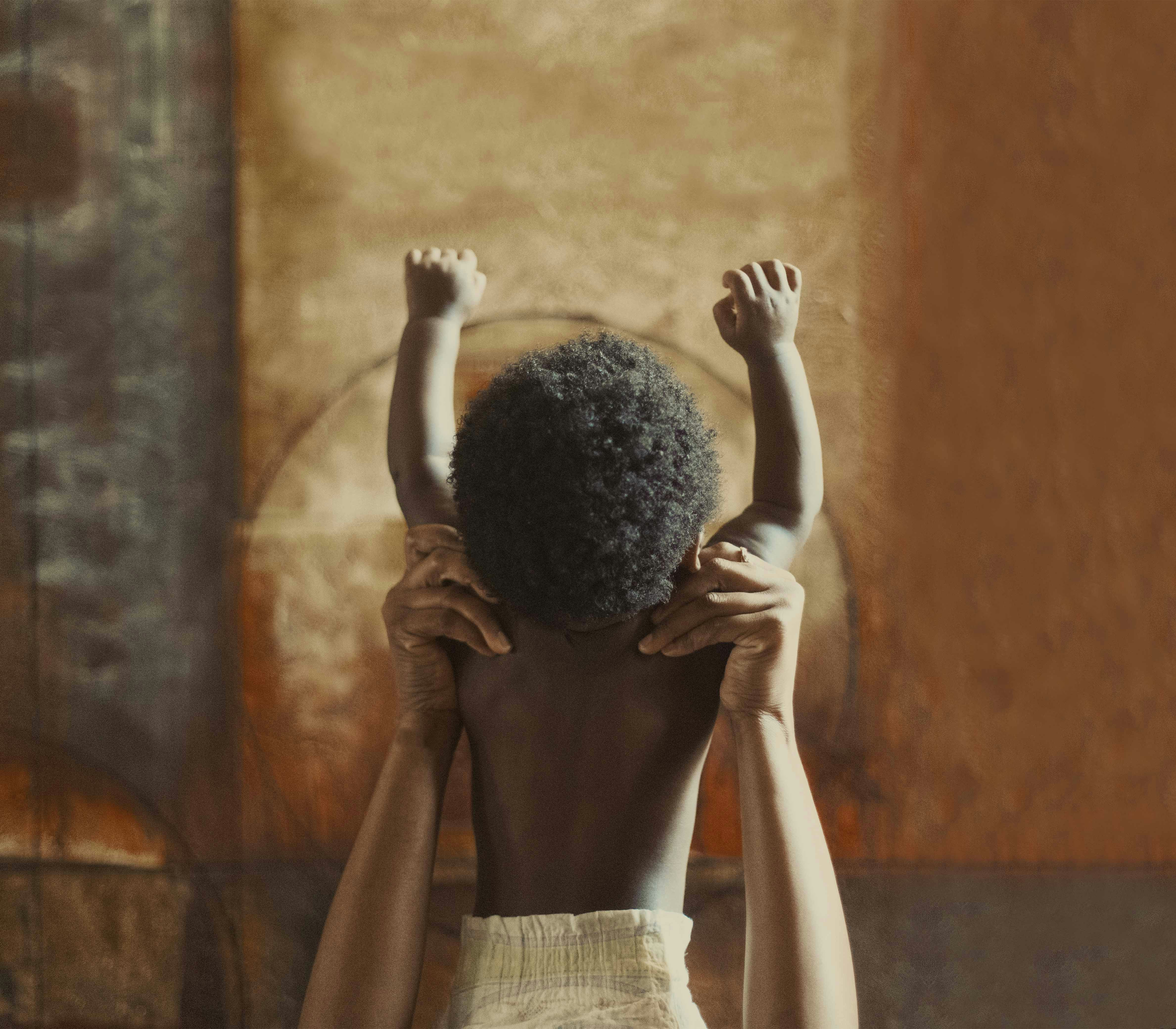 African baby with arms raised in studio. Adult holds baby. Warm color grading with brown and beige background and soft light.