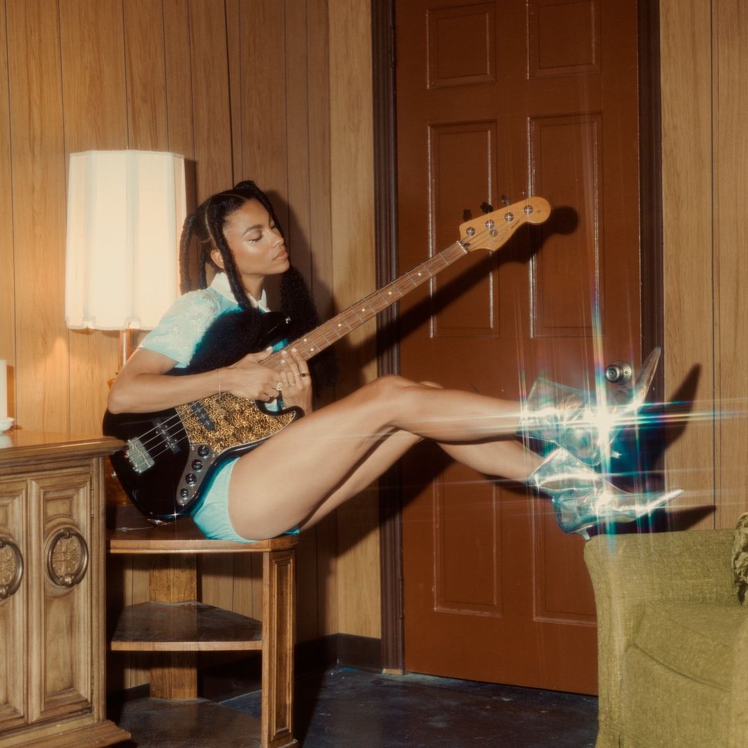 Adult female musician plays black bass guitar while sitting on a wood table in a room with wood paneled walls.
