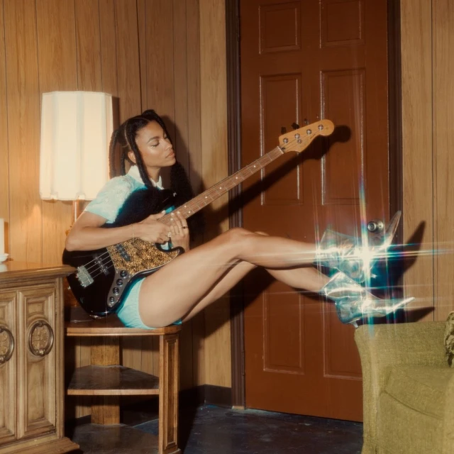 Adult female musician plays black bass guitar while sitting on a wood table in a room with wood paneled walls.