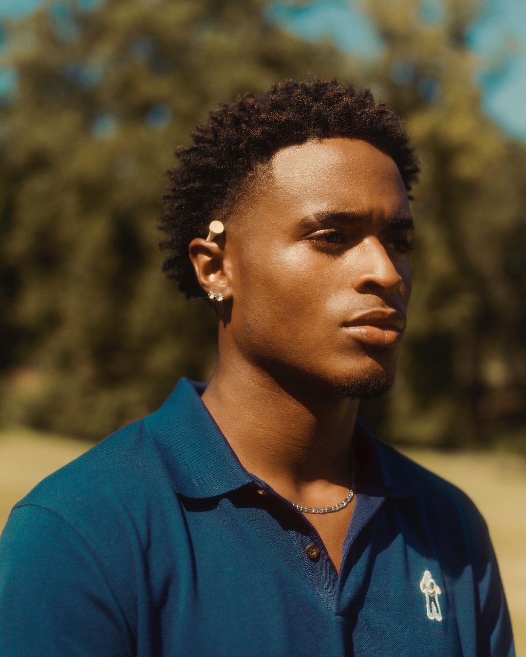 Close-up portrait of a young Black male in a blue polo shirt, featuring a shallow depth of field and a natural outdoor setting.