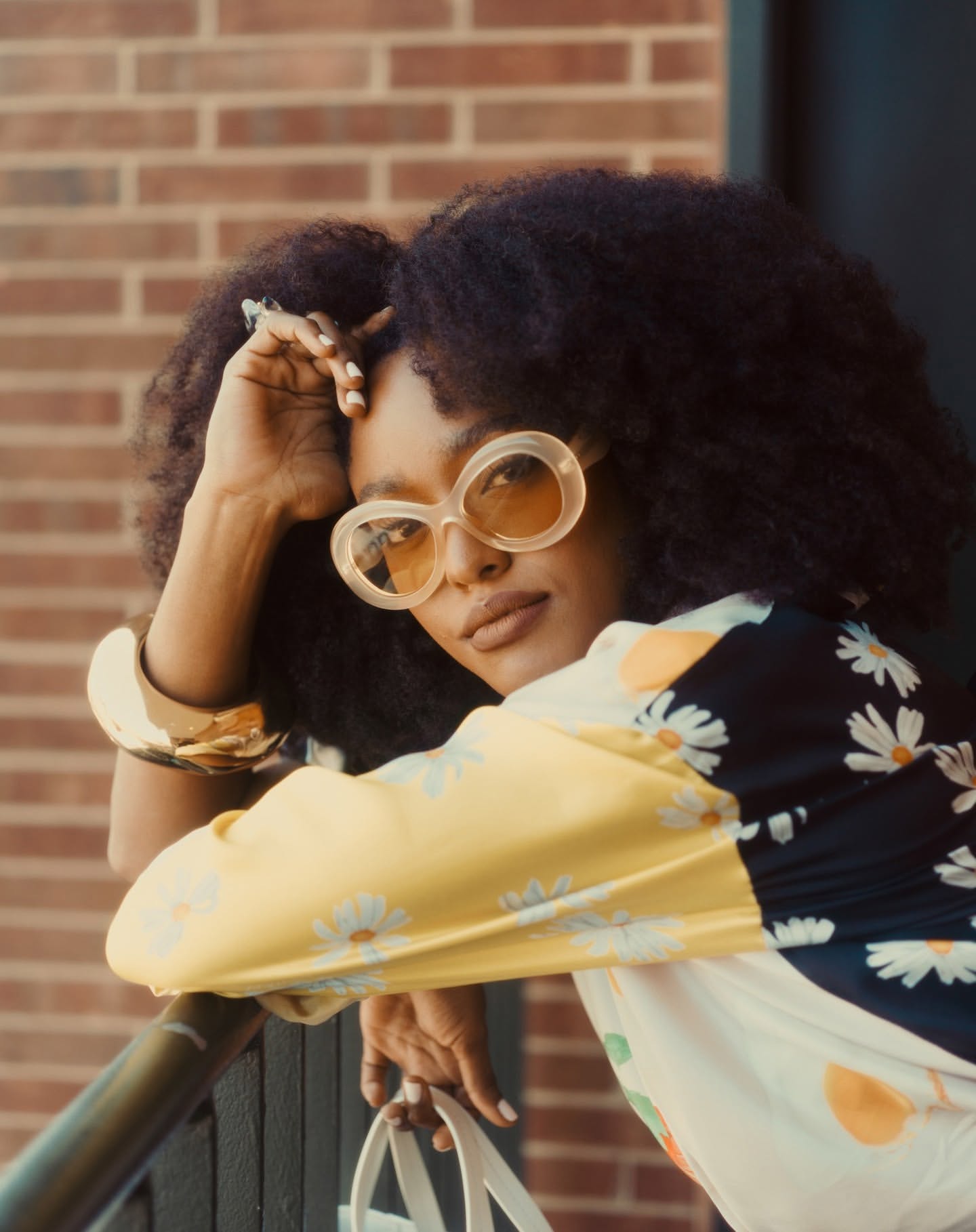Stylish woman with afro in retro sunglasses and floral top poses against a brick wall, accessorized with a gold bracelet and white tote.
