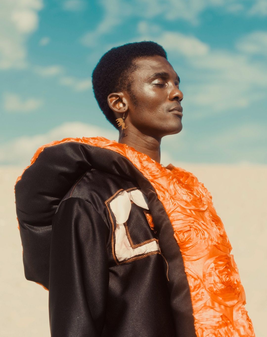 Black man in profile wearing a dark jacket and textured orange collar against a bright blue sky with clouds