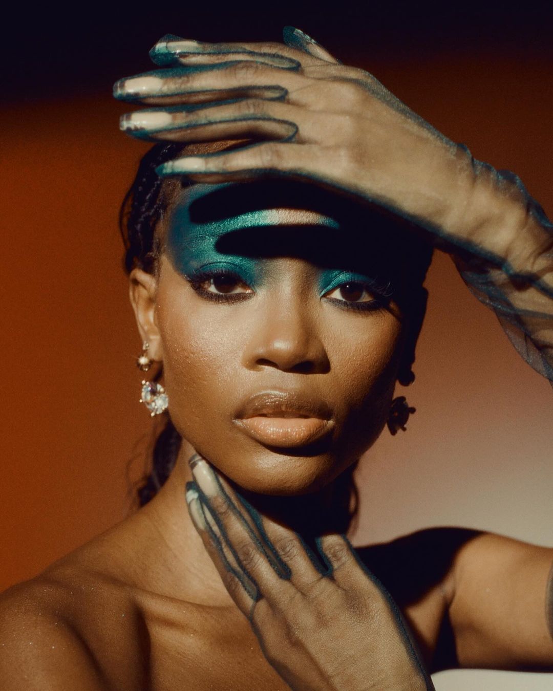 Close-up portrait of a woman with dark skin, styled with teal eyeshadow and sheer black gloves, set against an orange and brown gradient backdrop.