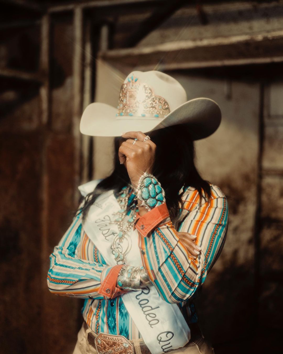 Portrait of a rodeo queen partially concealing her face with a decorated cowboy hat, showcasing turquoise jewelry and a striped shirt indoors.