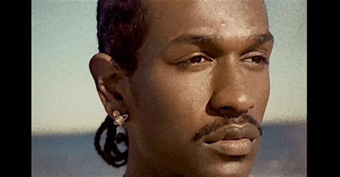 Close-up shot of a man with an earring, styled hair, and mustache against a bright background of the sky.