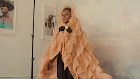 Fashionable Black woman poses in ruffled peach gown with black underdress, photo studio setting with framed prints on wall.