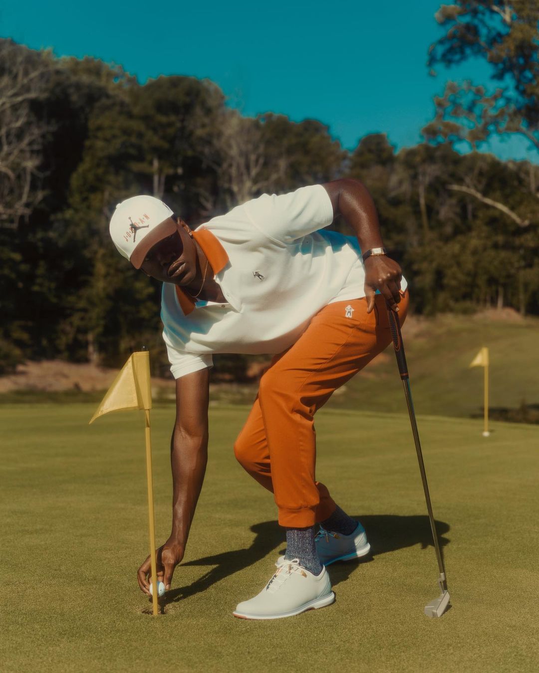 An African American man places a golf ball on a tee on a green golf course with a golf club nearby under a clear blue sky.