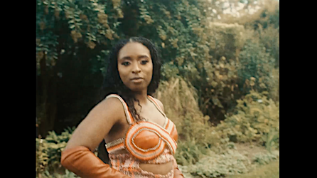 An adult female with long dark hair poses in an orange outfit with arm coverings in an outdoor environment with green foliage.