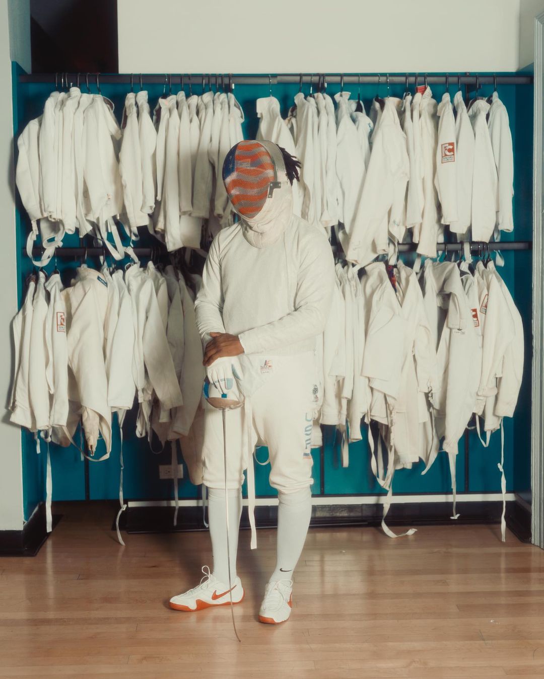 Fencer in white uniform and mask stands in front of a rack of white jackets in an indoor setting with a light brown floor.