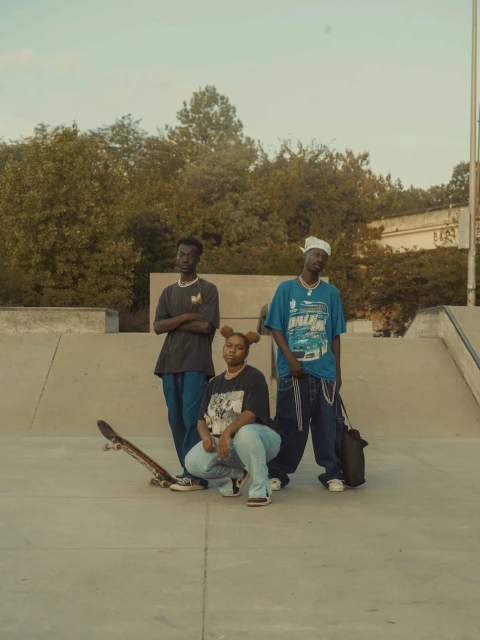 Three people pose at a skate park on a sunny day. Two young adult males stand while a young adult female crouches in front.