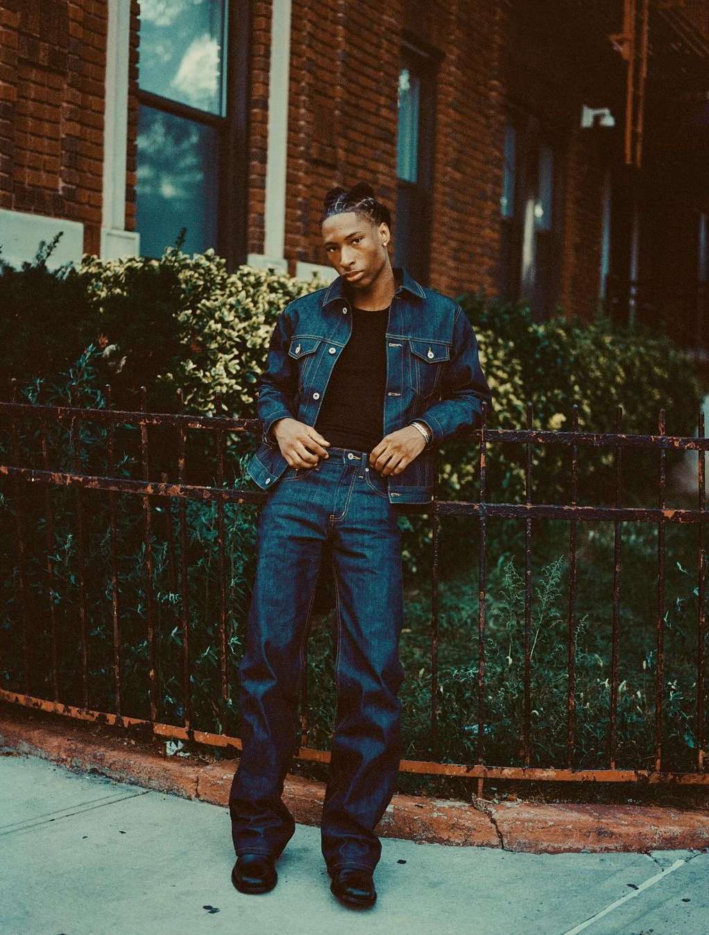 Stylish young adult man in denim jacket and jeans poses against brick building on sidewalk with green foliage in the background.