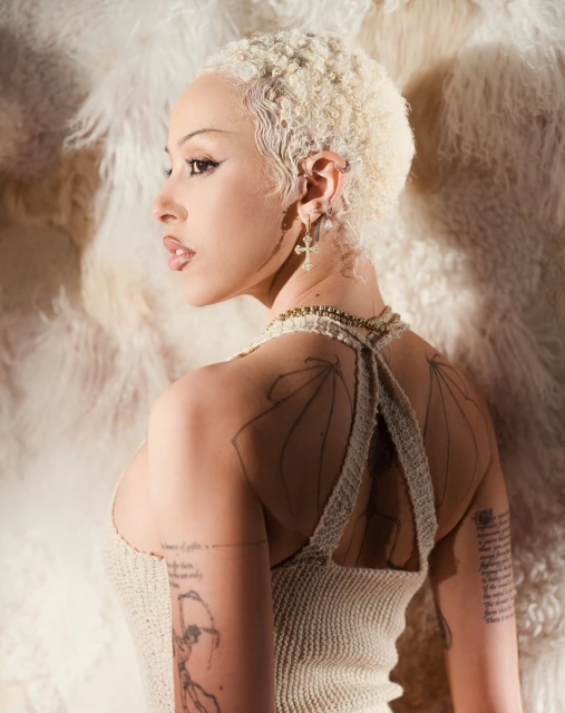Profile shot of a blonde woman with tattoos on her arms and back posing against a faux fur background, wearing a knit top.