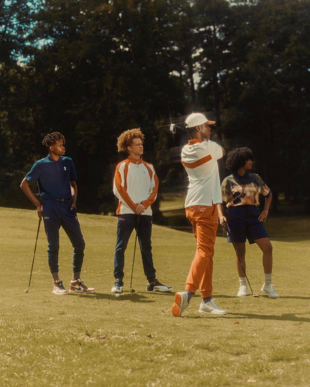 Four young adults are standing on a grassy golf course with trees in the background, one swinging a golf club in sunlight.