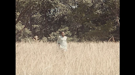 An adult woman in a light-colored dress walks through a field of wheat-like grass under the sun, with a forest visible in the background.