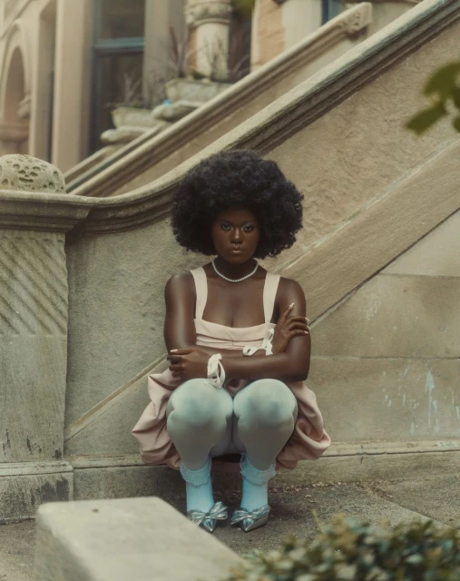 Portrait of a young Black woman squatting on stone steps, wearing a light pink dress, pearl necklace, and light blue shoes with bows.