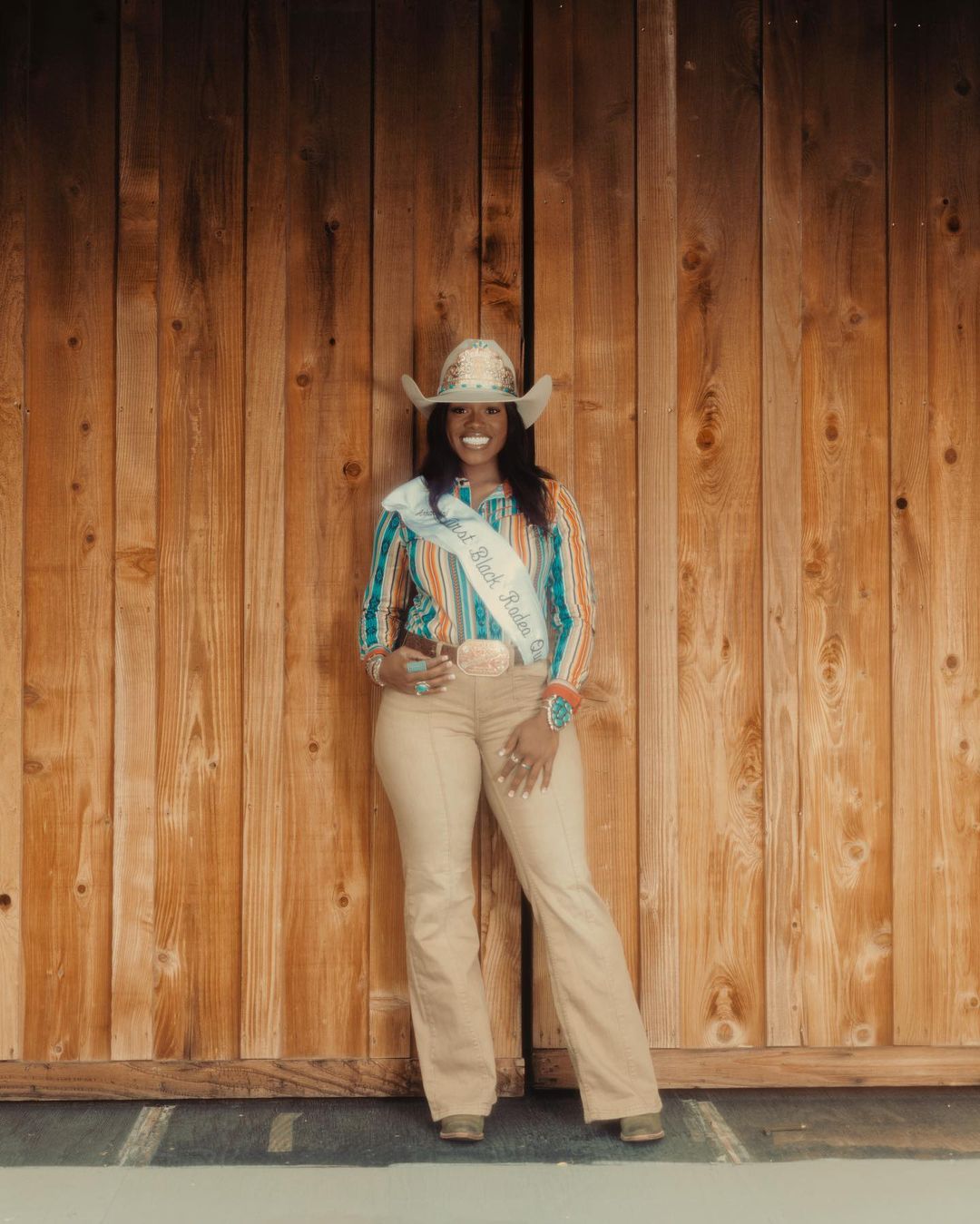 Smiling woman in cowboy hat and western wear poses in front of rustic wood wall, wearing a rodeo queen sash for a portrait.