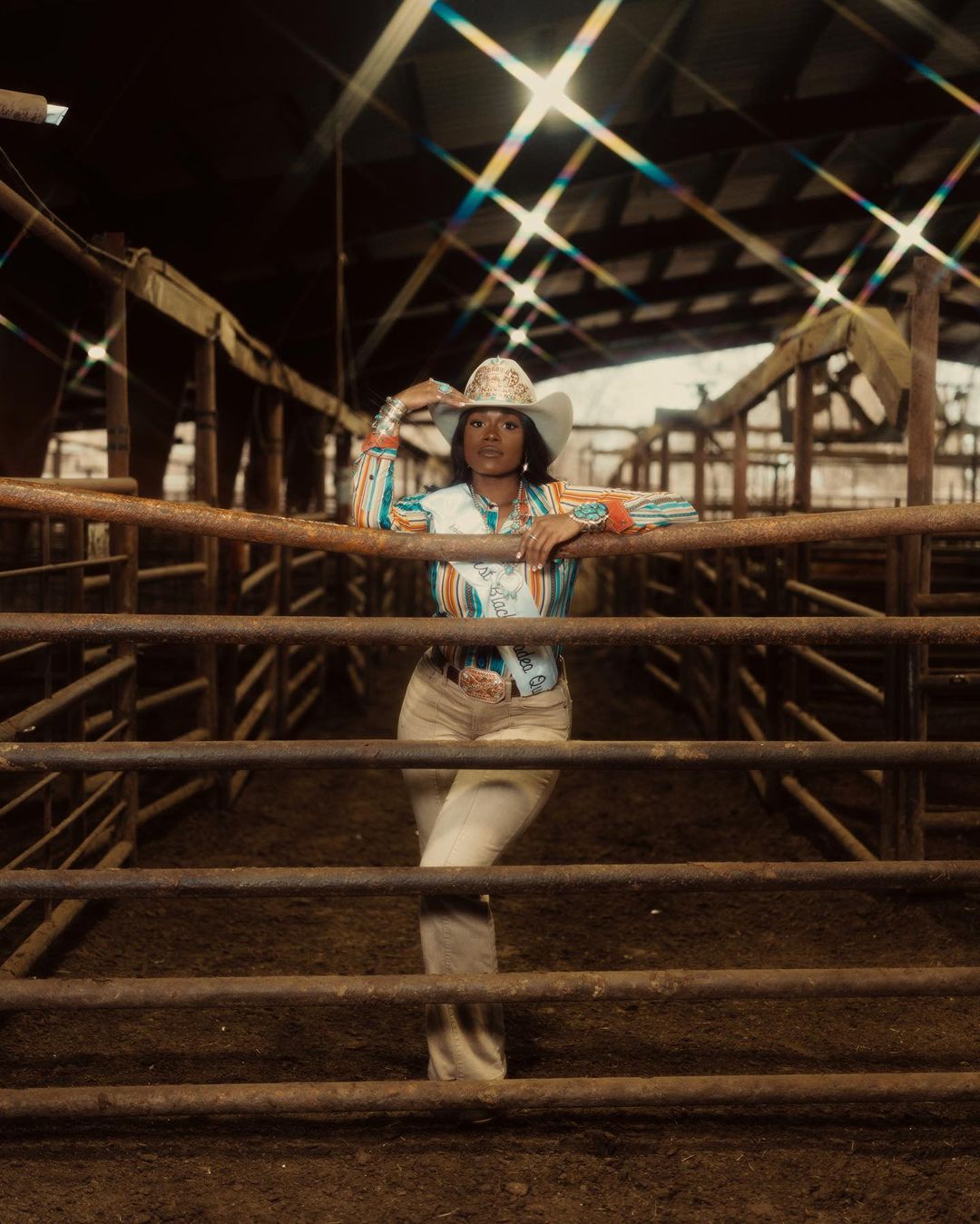 A woman in a cowboy hat poses indoors in an equestrian arena, wearing a colorful western shirt, jewelry, and jeans near a metal gate.