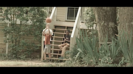 Young boy bounces a basketball outside as another boy sits on the porch stairs in a daytime shot with green foliage.