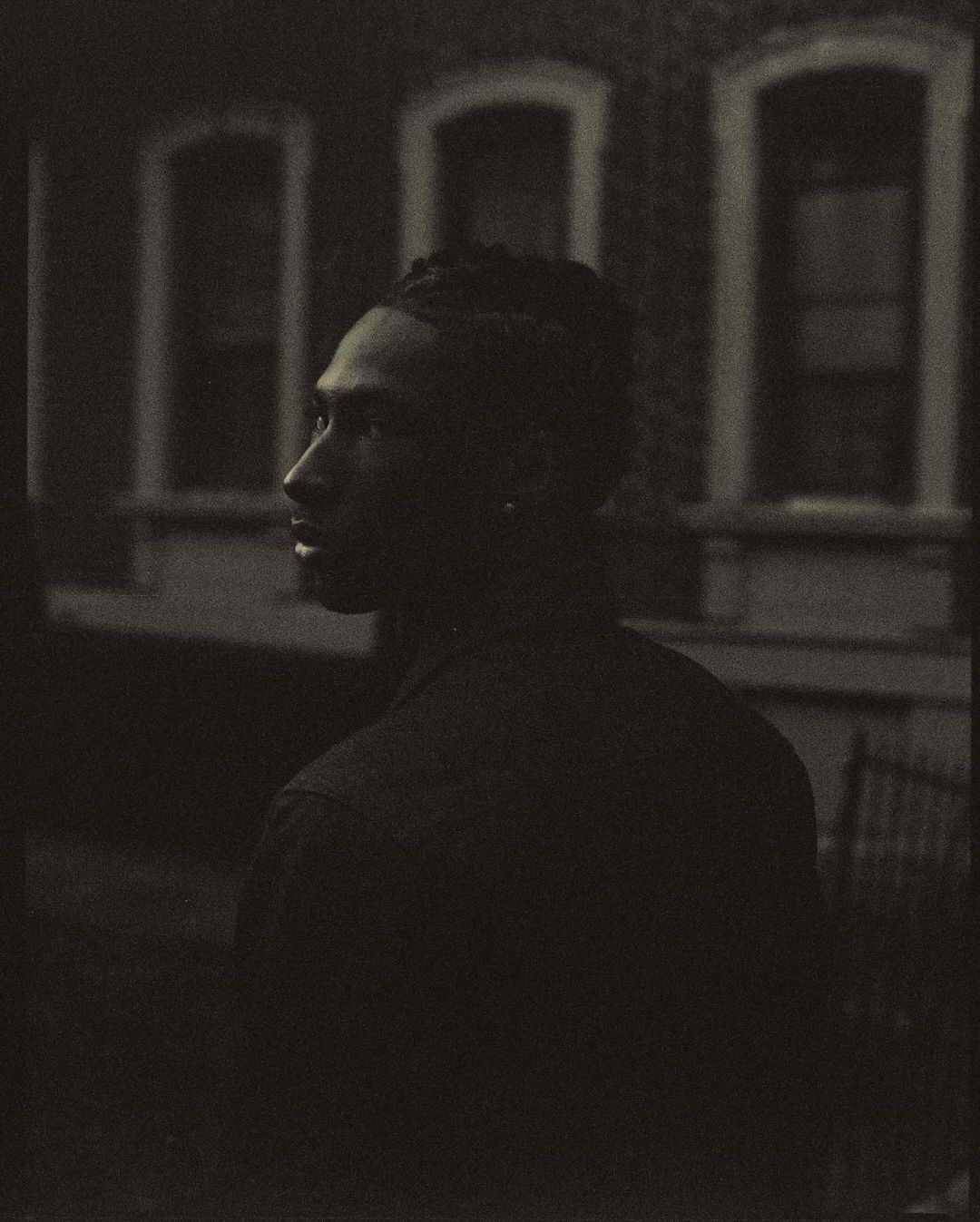 Monochrome portrait of a stylish adult male with locs, gazing thoughtfully with an architectural backdrop featuring framed windows.