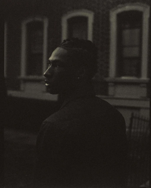 Monochrome portrait of a stylish adult male with locs, gazing thoughtfully with an architectural backdrop featuring framed windows.